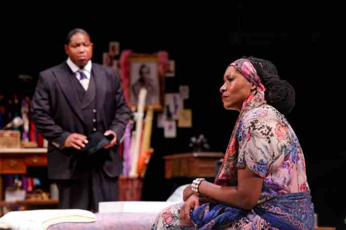 A Black woman wearing a floral dress and scarf sits while a Black man looks at her.